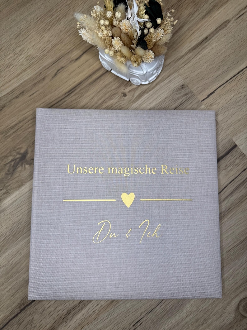 Op de afbeelding: Een vierkant fotoalbum met een lichtgrijze linnen omslag. De voorzijde is voorzien van goudfolie tekst met "Unsere magische Reise" boven een gouden hartpictogram en de tekst "Du & Ich" in een cursief lettertype.
