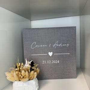 Op de afbeelding: Een vierkante, met grijze stof beklede doos met de witte tekst Corinna & Andreas, een klein hartje en de datum 21.12.2024. Daarnaast staat een witte keramische plantenbak in de vorm van een busje, gevuld met gedroogde beige en zwarte grassen.