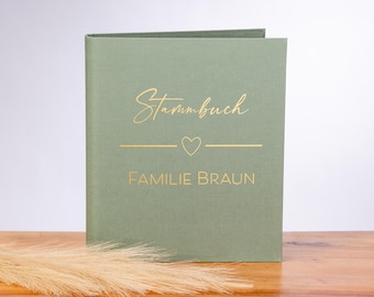 Op de afbeelding: Een saliegroen boek met gouden letters met de tekst "Stammbuch" boven een hartsymbool en "Familie Braun". Het boek ligt op een houten oppervlak met gedroogd siergras op de voorgrond. De kaft van het boek staat iets open.