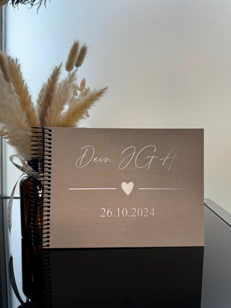 Op de afbeelding: Een spiraalgebonden notitieboek met een lichtbeige, stoffen omslag. De voorkant bevat de zilverkleurige tekst "Dein JGA" boven een klein hartje en een horizontale lijn, met de datum "26.10.2024" in zilverkleur onder het hartje gedrukt.