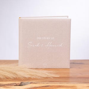 Op de afbeelding: Een beige linnen fotoalbum met de tekst "The Story of Sarah & Heinrich" in wit schrift en een klein hartje.