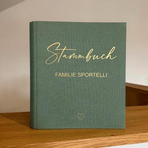 Könnte beinhalten: Ein grünes Hardcover-Buch mit goldener Schrift. Auf dem Einband steht "Stammbuch" und "Familie Sportelli." Unten befindet sich ein kleines Herz. Das Buch liegt auf einem Holzregal.