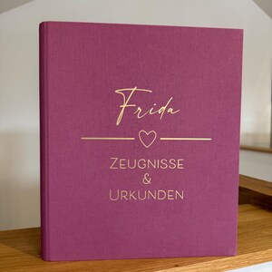 Könnte beinhalten: Ein Hardcover-Buch mit magenta Einband. Auf dem Einband steht in goldener Schrift der Name "Frida", darunter eine goldene Linie mit Herz und die Worte "ZEUGNISSE & URKUNDEN" in Gold. Das Buch liegt auf einem Holzregal.