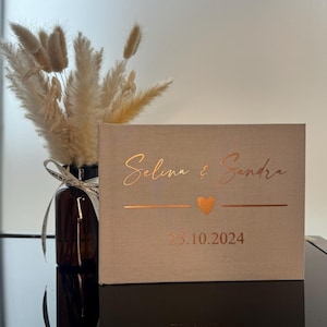Op de afbeelding: Een rechthoekig fotoalbum bekleed met beige stof, voorzien van koperkleurige folietekst met de namen Selina & Sandra, een klein hartje en de datum 25.10.2024. Het album heeft een strak ontwerp met een gestructureerde, linnenachtige afwerking.