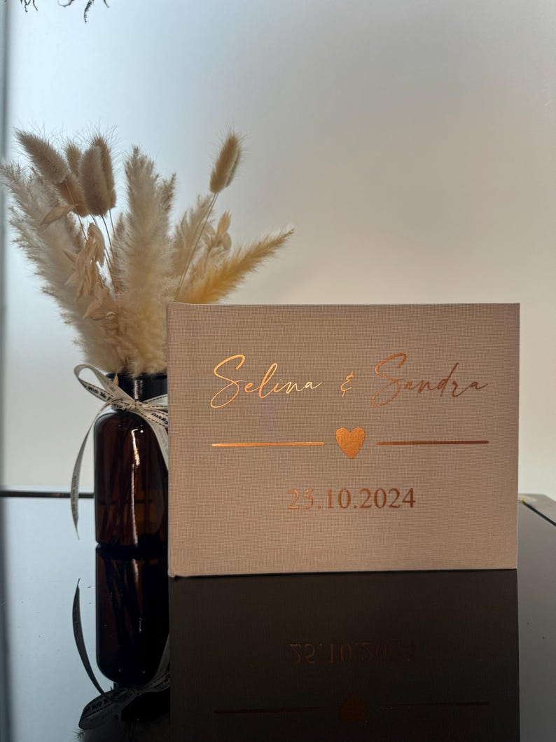 Op de afbeelding: Een rechthoekig fotoalbum bekleed met beige stof, voorzien van koperkleurige folietekst met de namen Selina & Sandra, een klein hartje en de datum 25.10.2024. Het album heeft een strak ontwerp met een gestructureerde, linnenachtige afwerking.