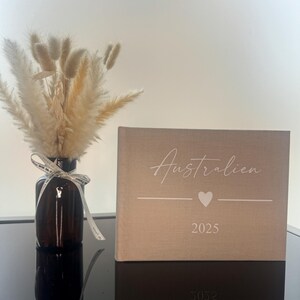 Op de afbeelding: Een beige met stof bekleed fotoalbum met de tekst "Australien", een hartpictogram en "2025" in het wit gedrukt. Daarnaast staat een donkerbruine glazen fles met een boeket van gedroogd beige pampasgras en lagurus-stengels.