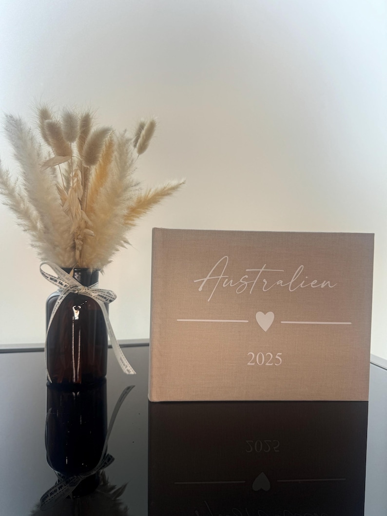 Op de afbeelding: Een beige met stof bekleed fotoalbum met de tekst "Australien", een hartpictogram en "2025" in het wit gedrukt. Daarnaast staat een donkerbruine glazen fles met een boeket van gedroogd beige pampasgras en lagurus-stengels.