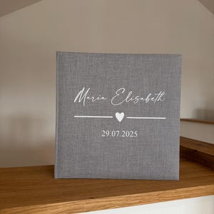 Op de afbeelding: Een vierkant fotoalbum met een grijze stoffen kaft. De voorzijde is voorzien van de wit gedrukte tekst "Maria Elisabeth" in een schrijflettertype, gevolgd door een horizontale lijn met een klein hartje in het midden en de datum "29.07.2025".