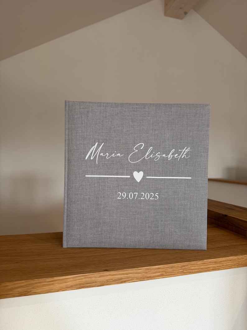 Op de afbeelding: Een vierkant fotoalbum met een grijze stoffen kaft. De voorzijde is voorzien van de wit gedrukte tekst "Maria Elisabeth" in een schrijflettertype, gevolgd door een horizontale lijn met een klein hartje in het midden en de datum "29.07.2025".