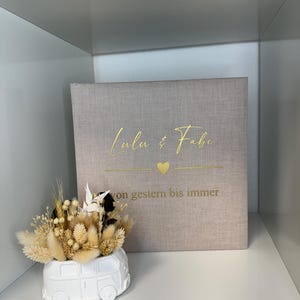 Op de afbeelding: Een vierkant, lichtbeige met stof bekleed fotoalbum met goudkleurige tekst Lulu & Fabi, een klein hartje en de tekst von gestern bis immer. Daarnaast staat een witte keramische plantenbak in de vorm van een busje, gevuld met droogbloemen.