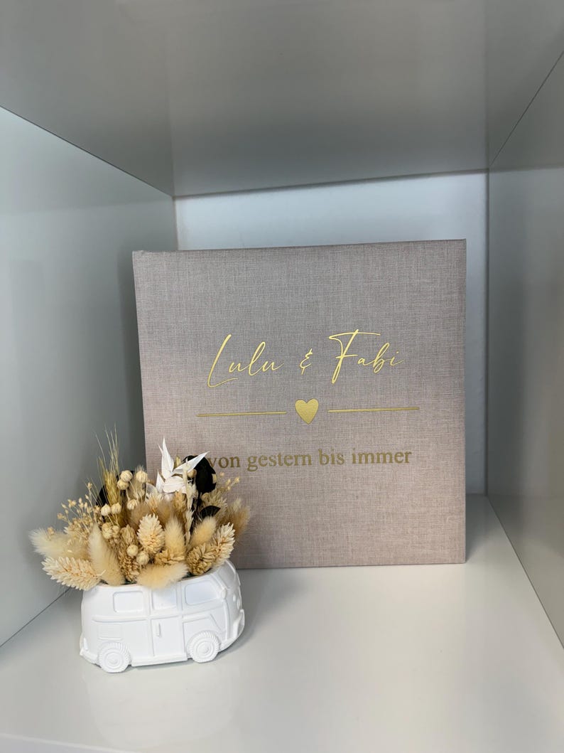 Op de afbeelding: Een vierkant, lichtbeige met stof bekleed fotoalbum met goudkleurige tekst Lulu & Fabi, een klein hartje en de tekst von gestern bis immer. Daarnaast staat een witte keramische plantenbak in de vorm van een busje, gevuld met droogbloemen.