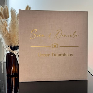 Op de afbeelding: Een vierkant, beige, met stof bekleed boek of doos met goudfolie belettering die "Sven & Daniela" leest boven een klein huisicoon met een hartje erin, gevolgd door de tekst "Unser Traumhaus" in een strak, schreefloos lettertype.
