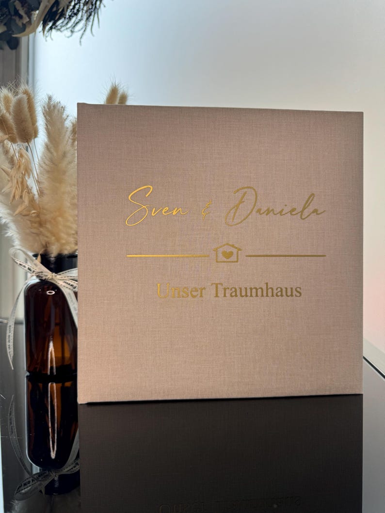 Op de afbeelding: Een vierkant, beige, met stof bekleed boek of doos met goudfolie belettering die "Sven & Daniela" leest boven een klein huisicoon met een hartje erin, gevolgd door de tekst "Unser Traumhaus" in een strak, schreefloos lettertype.