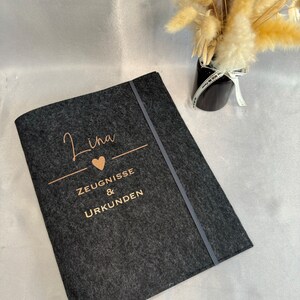 Könnte beinhalten: Ein schwarzer Filzordner mit einer goldenen Aufschrift, die "Lina" mit einem Herzsymbol, "Zeugnisse & Urkunden" lautet.