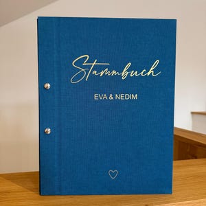Op de afbeelding: Een blauw hardcover boek met het woord "Stammbuch" in gouden script. De namen "EVA & NEDIM" staan onder de titel gedrukt, met een klein hartsymbool. Het boek ligt op een houten oppervlak.