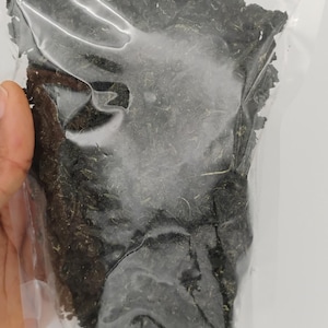 May include: A clear plastic bag filled with dark green, dried, leafy herbs. The bag is held in a person's hand against a white background. The herbs appear to be finely chopped and have a slightly rough texture.
