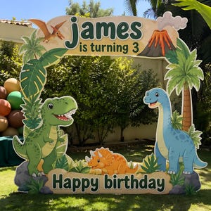 Può includere: Un supporto per fototessera a tema dinosauri con la scritta "James compie 3 anni" e "Buon compleanno". Il design presenta dinosauri dei cartoni animati, un vulcano, palme e uno pterodattilo volante, perfetto per una festa di compleanno.