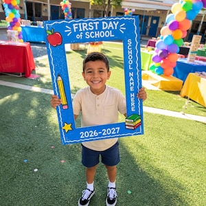 May include: A blue rectangular photo frame prop with white text reading FIRST DAY of SCHOOL and 2026-2027. The frame features illustrations of a red apple, a yellow pencil, a yellow star, and a stack of colorful books, with space for a school name.