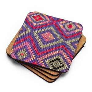 May include: Set of four square coasters with a colorful, crocheted design. The pattern features a repeating geometric motif in shades of purple, pink, red, and beige. The coasters have a cork backing.