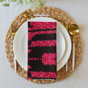 May include: A folded black napkin featuring a striking pink and orange geometric design, set on a white plate with gold cutlery. The table setting includes a woven placemat and a neutral tablecloth, creating an elegant dining scene.