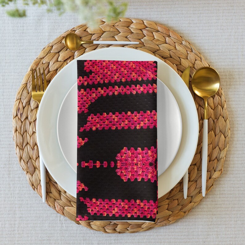 May include: A folded black napkin featuring a striking pink and orange geometric design, set on a white plate with gold cutlery. The table setting includes a woven placemat and a neutral tablecloth, creating an elegant dining scene.