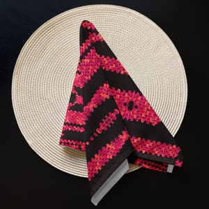 May include: A folded black napkin with a vibrant pink and red geometric pattern, placed on a beige woven placemat. The napkin's design features a repeating, textured pattern. The background is a dark surface.