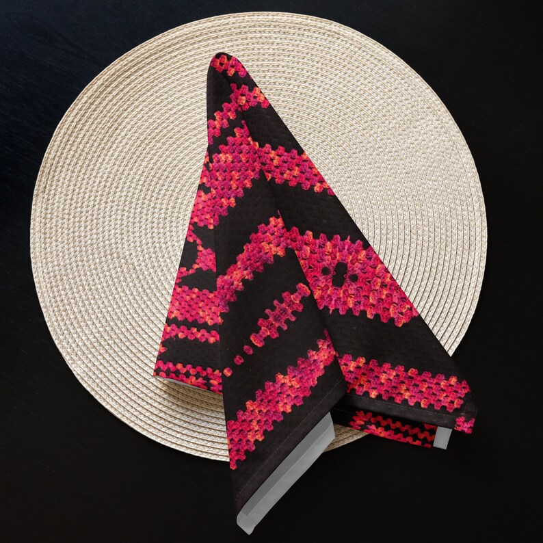 May include: A folded black napkin with a vibrant pink and red geometric pattern, placed on a beige woven placemat. The napkin's design features a repeating, textured pattern. The background is a dark surface.