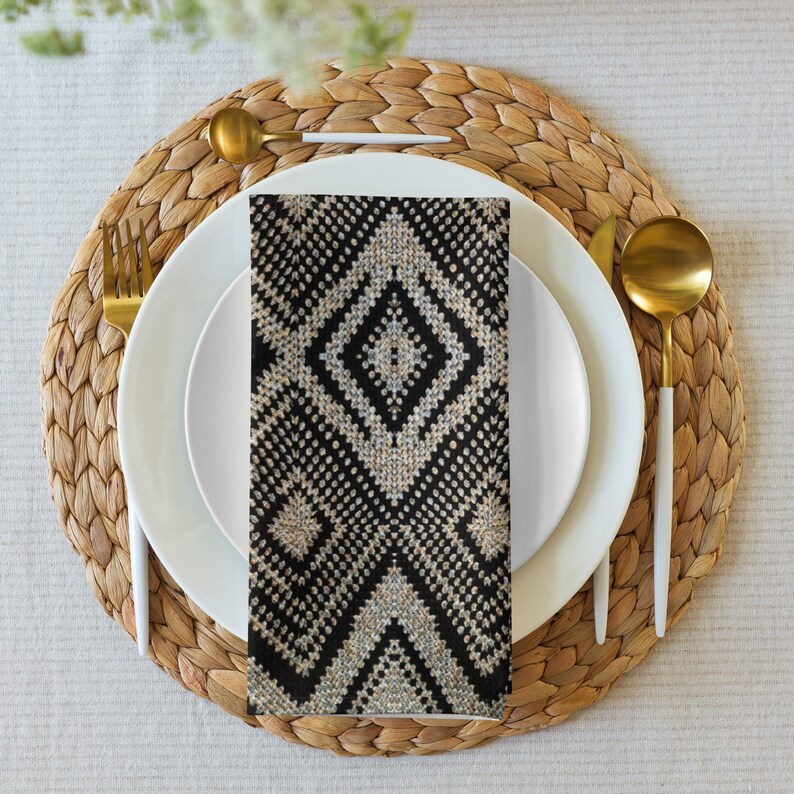 May include: A black and white patterned napkin is placed on a white plate, with gold-colored silverware and a woven placemat. The napkin features a geometric diamond design. The table setting is on a neutral-colored tablecloth.