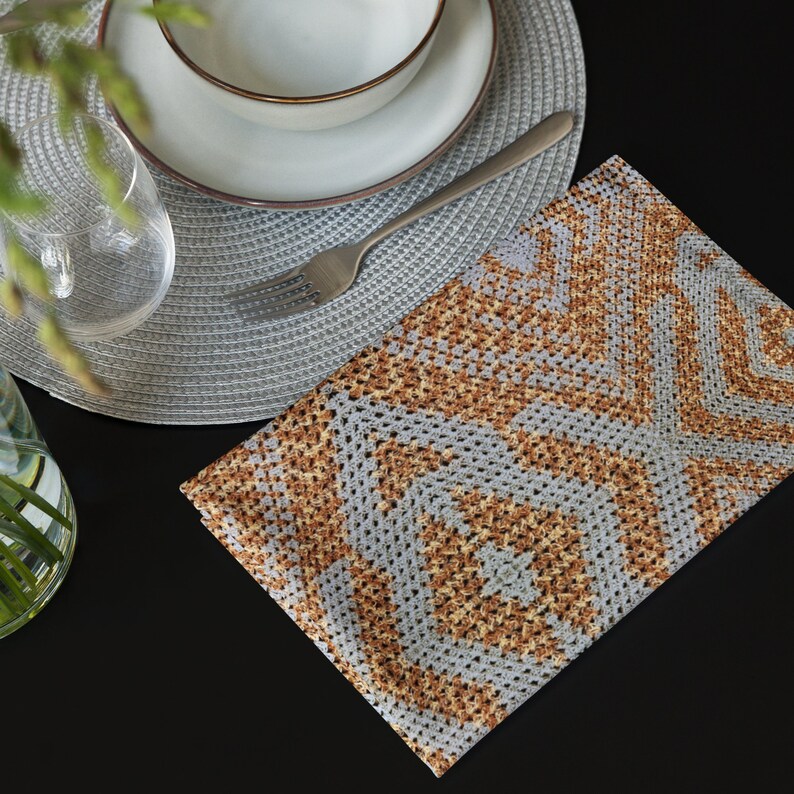 May include: A grey and brown crocheted napkin with a geometric pattern on a black surface. A white bowl with a gold rim, a plate, a fork, and a glass are also visible, suggesting a dining setting.