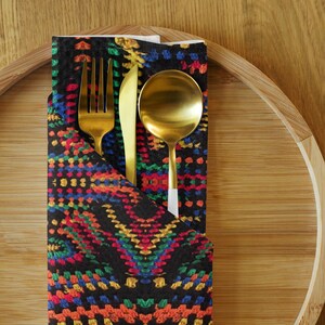 May include: A gold-colored fork, knife, and spoon set inside a black napkin with a colorful, geometric pattern. The napkin is placed on a bamboo serving tray, creating a stylish table setting.