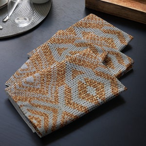 May include: Three folded square napkins with a geometric pattern in shades of grey and brown. The napkins are made of a textured fabric and are arranged on a dark surface, suggesting a dining setting.
