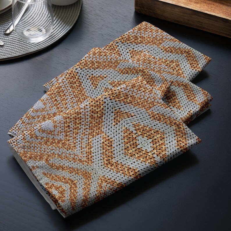 May include: Three folded square napkins with a geometric pattern in shades of grey and brown. The napkins are made of a textured fabric and are arranged on a dark surface, suggesting a dining setting.