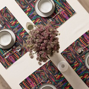 May include: Overhead shot of a table setting with four placemats. The placemats have a black background with a colorful, geometric pattern. Each place setting includes a white plate, bowl, silverware, and a small vase. A decorative plant is in the center.