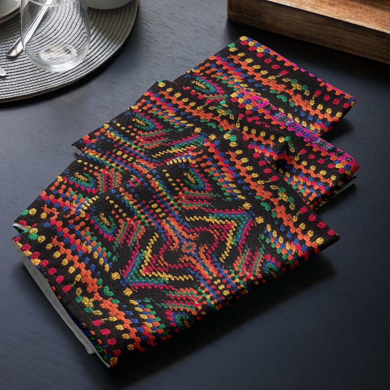 Op de afbeelding: Drie gevouwen zwarte stoffen servetten met een levendig, geometrisch patroon in rood, oranje, geel, groen en blauw. De servetten zijn op een donkere ondergrond gestapeld en tonen het kleurrijke ontwerp.