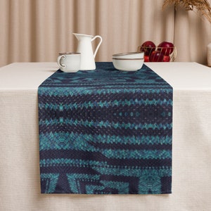 Puede incluir: Un camino de mesa azul oscuro con un patr&oacute;n geom&eacute;trico turquesa. El camino de mesa se muestra sobre un mantel blanco, con una jarra, taza y cuencos blancos en el fondo. Una cesta de alambre dorada contiene manzanas rojas.