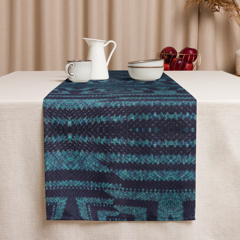 Puede incluir: Un camino de mesa azul oscuro con un patr&oacute;n geom&eacute;trico turquesa. El camino de mesa se muestra sobre un mantel blanco, con una jarra, taza y cuencos blancos en el fondo. Una cesta de alambre dorada contiene manzanas rojas.