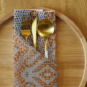 May include: A gold-coloured fork, knife, and spoon set rests on a folded napkin with a geometric pattern in shades of grey and brown. The cutlery is arranged on a bamboo serving tray, creating an elegant table setting.