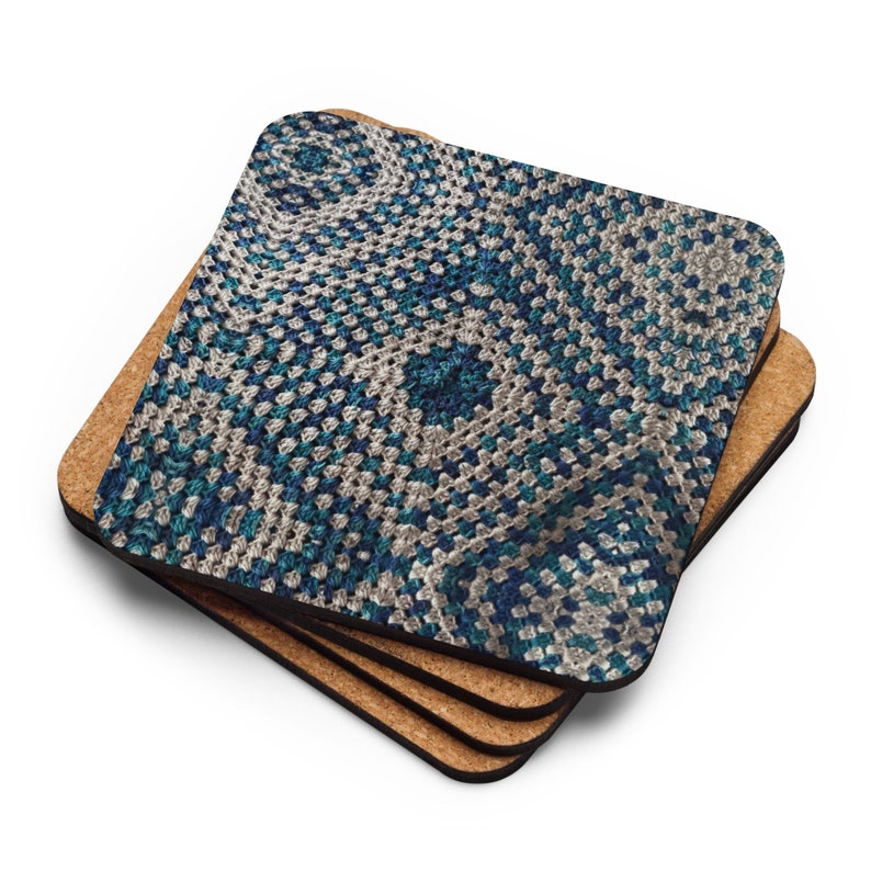 Op de afbeelding: Een stapel van vier vierkante onderzetters. De bovenste onderzetter heeft een gedetailleerd haakpatroon in tinten blauw, teal en wit. De onderzetters hebben een kurken achterkant. Het haakontwerp is een herhalend geometrisch patroon.