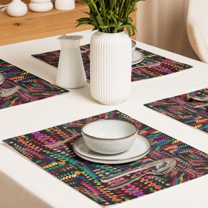 May include: A dining table setting with four colorful placemats featuring a vibrant, geometric pattern in shades of red, yellow, green, and blue on a black background. The table is set with white dishes, silverware, and a white vase with green foliage.
