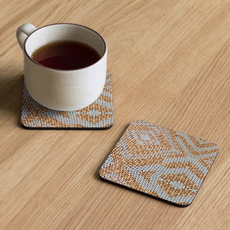 May include: Two square coasters with a geometric pattern in shades of tan, grey, and white. One coaster has a white mug with tea on top. The coasters are on a light wood surface.