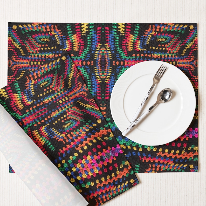 May include: A black placemat and napkin set with a vibrant, colorful pattern. The design features a repeating, abstract motif in shades of red, yellow, green, and blue. A white plate, fork, and spoon are on the placemat.
