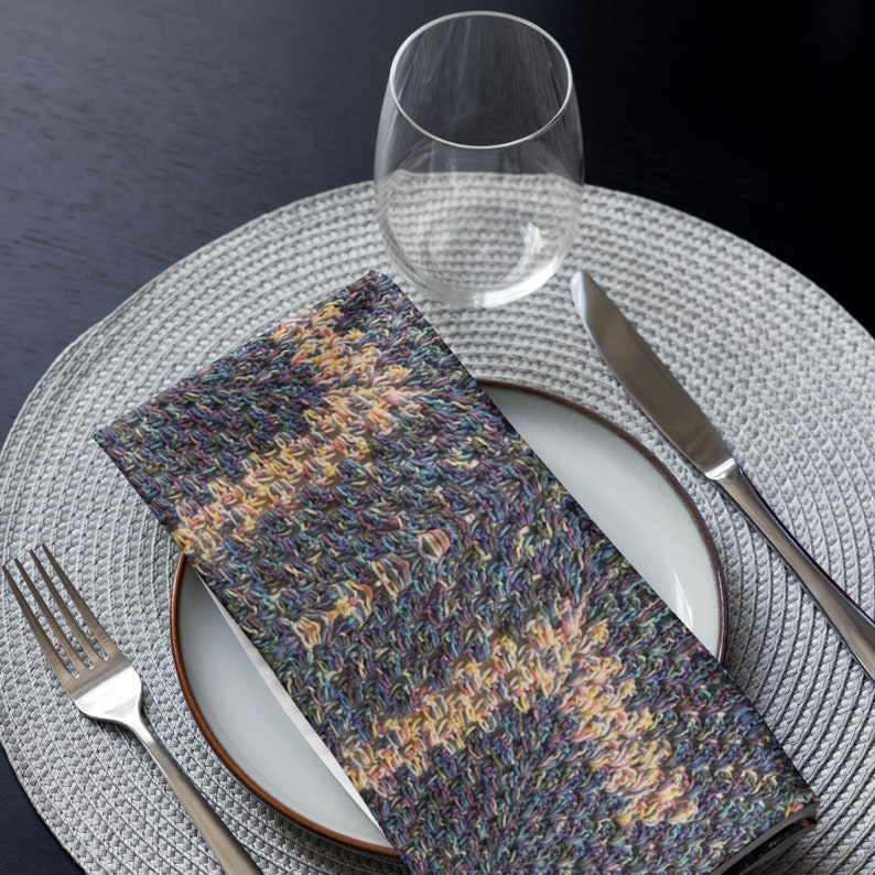 Puede incluir: Una servilleta doblada con un patr&oacute;n colorido y texturizado descansa sobre un plato blanco. Una copa de vino, un tenedor y un cuchillo est&aacute;n dispuestos sobre un mantel individual gris, creando un ambiente formal.