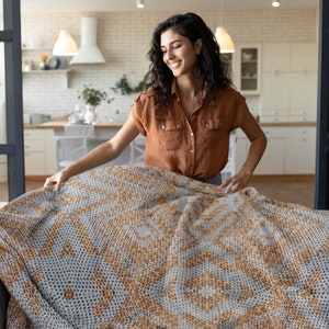 Pode incluir: Uma mulher a arrumar uma manta grande estampada. A manta apresenta um design geom&eacute;trico repetitivo em tons de bege e cinza. A mulher est&aacute; a usar uma camisa castanha abotoada.