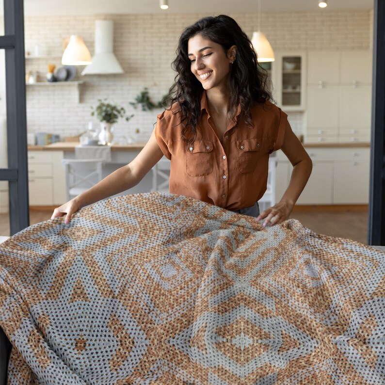 Pode incluir: Uma mulher a arrumar uma manta grande estampada. A manta apresenta um design geom&eacute;trico repetitivo em tons de bege e cinza. A mulher est&aacute; a usar uma camisa castanha abotoada.