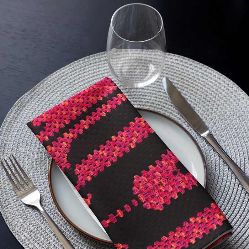 May include: A folded black and pink patterned napkin rests on a white plate, next to a fork and knife. The napkin features a repeating floral design in shades of pink and orange. A clear wine glass sits in the background.