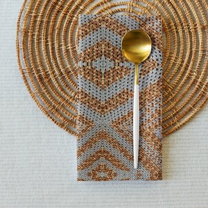 May include: A gold spoon with a white handle lies on a patterned napkin. The napkin has a geometric design in grey and brown tones. A woven placemat provides a textured backdrop, all set on a light-coloured surface.