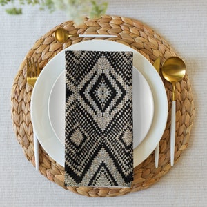 May include: A black and white patterned napkin is placed on a white plate, with gold-colored silverware and a woven placemat. The napkin features a geometric diamond design. The table setting is on a neutral-colored tablecloth.