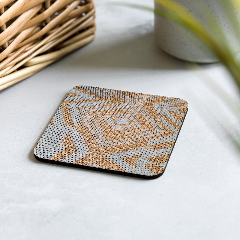 May include: Square coaster with a woven pattern in shades of tan and grey. The coaster has a black border and a textured surface. The design features a repeating geometric pattern. The coaster is designed to protect surfaces from drink rings.