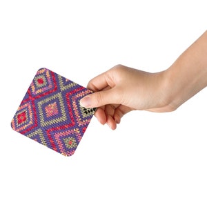 May include: A square coaster with a colorful geometric pattern. The design features diamond shapes in shades of purple, red, pink, and yellow. The coaster is held by a hand against a white background.