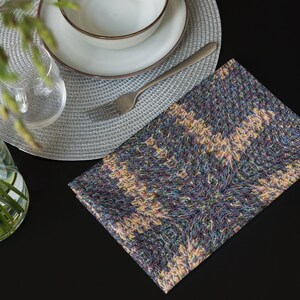 Puede incluir: Una servilleta de tela plegada, estampada y multicolor, est&aacute; sobre una superficie negra. Tambi&eacute;n se ven un cuenco y un plato blancos con borde marr&oacute;n, un tenedor plateado y un vaso transparente. La servilleta tiene un aspecto tejido y texturizado.